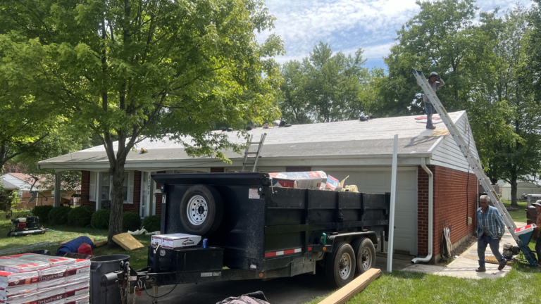 Brick ranch mid-re-roof with a loaded dump trailer and ladders against the eaves