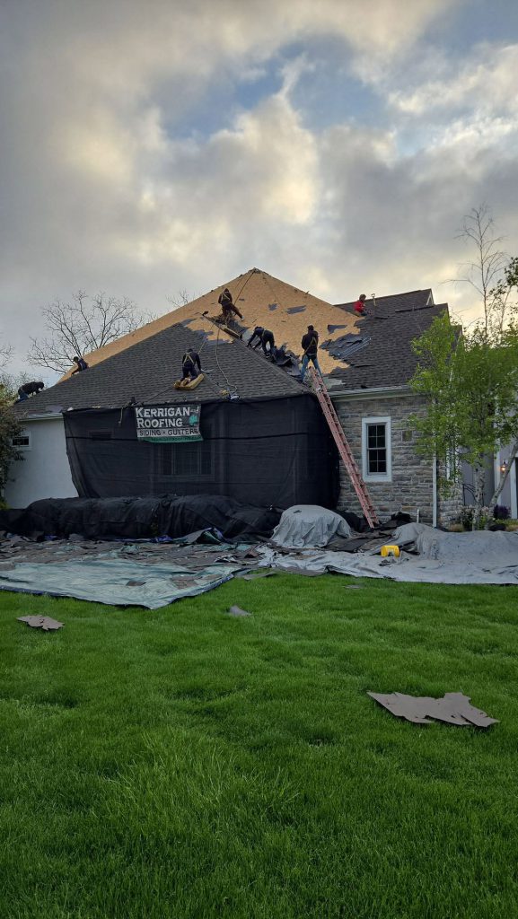 Kerrigan Roofing crew tearing off and replacing the shingle roof of a stone house, company banner hung on the protective sheeting