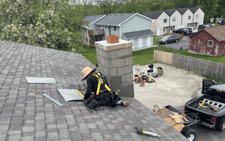Roofer in a safety harness working beside a chimney on a grey shingle roof