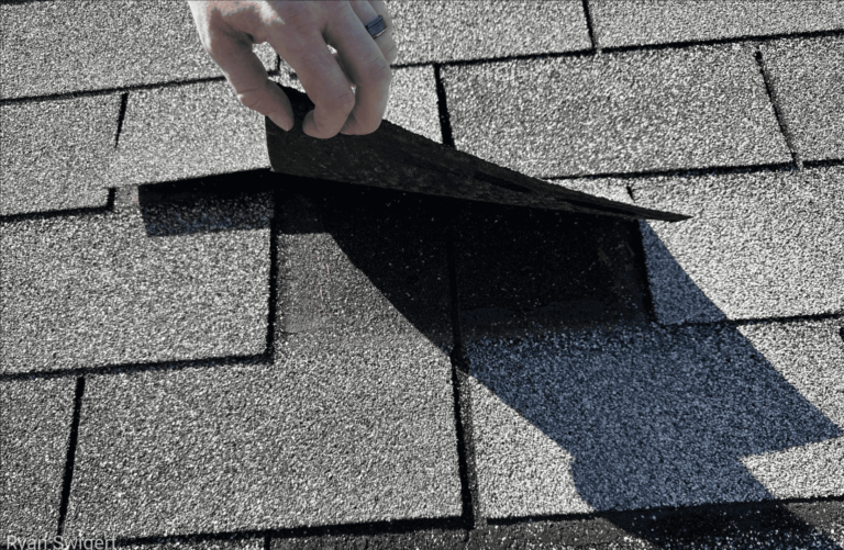 Hand lifting a cracked shingle tab to inspect the roof surface