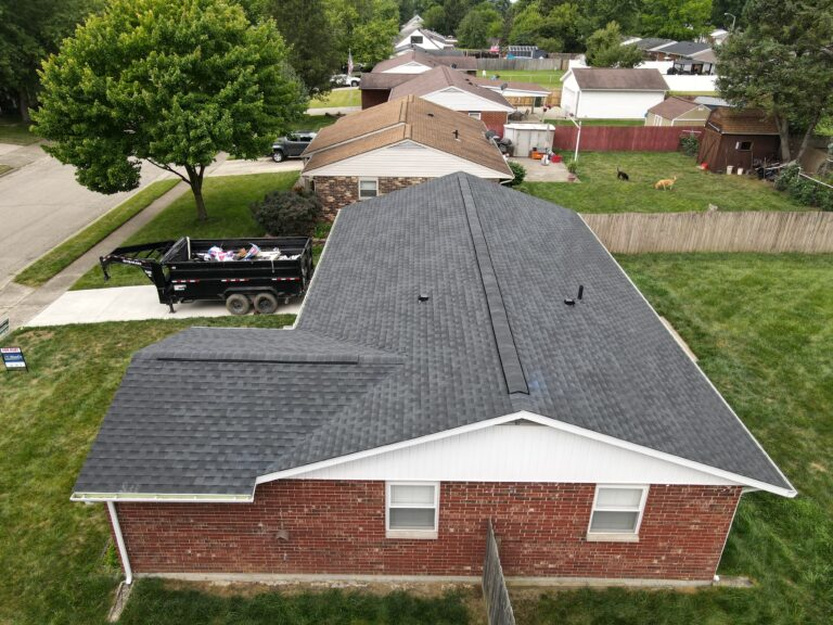 Aerial view of a brick ranch home with a new dark shingle roof and a dump trailer on the driveway