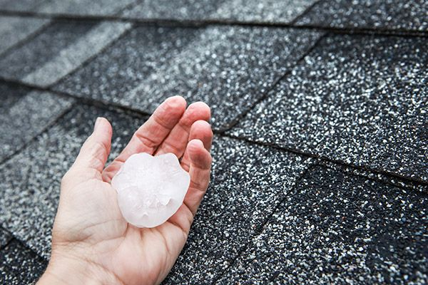Hand holding a large hailstone above a hail-battered asphalt shingle roof