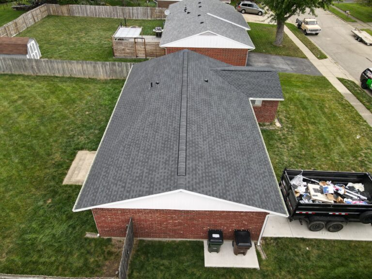Aerial view of a brick home with a new dark grey shingle roof beside a debris trailer