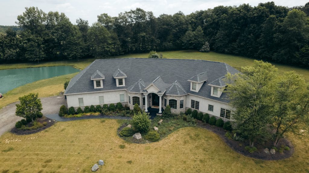 Aerial view of a large cream home with a grey shingle roof beside a pond